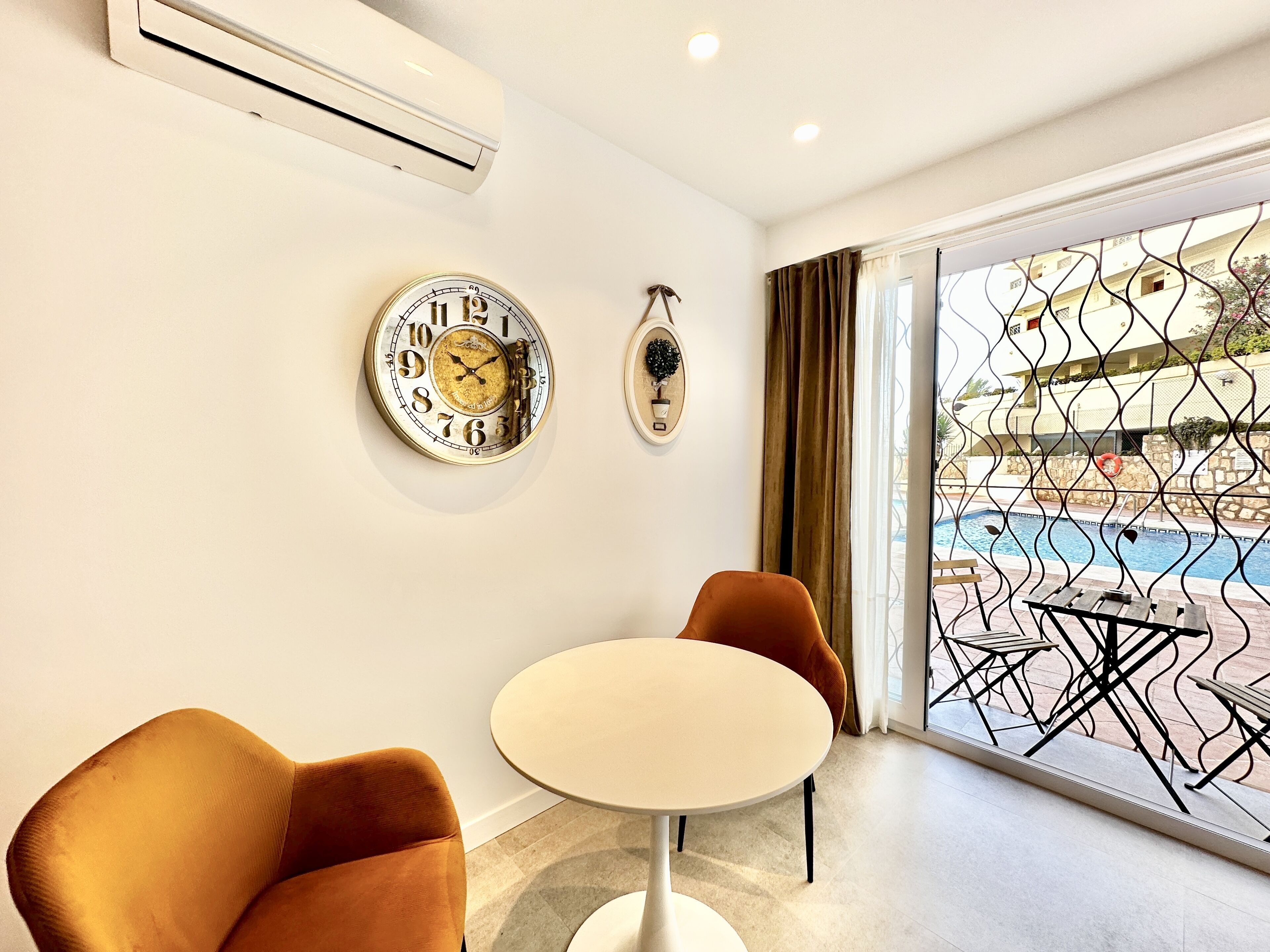 A modern room with a round table, two chairs, a wall clock, and a view of a pool through a glass door.