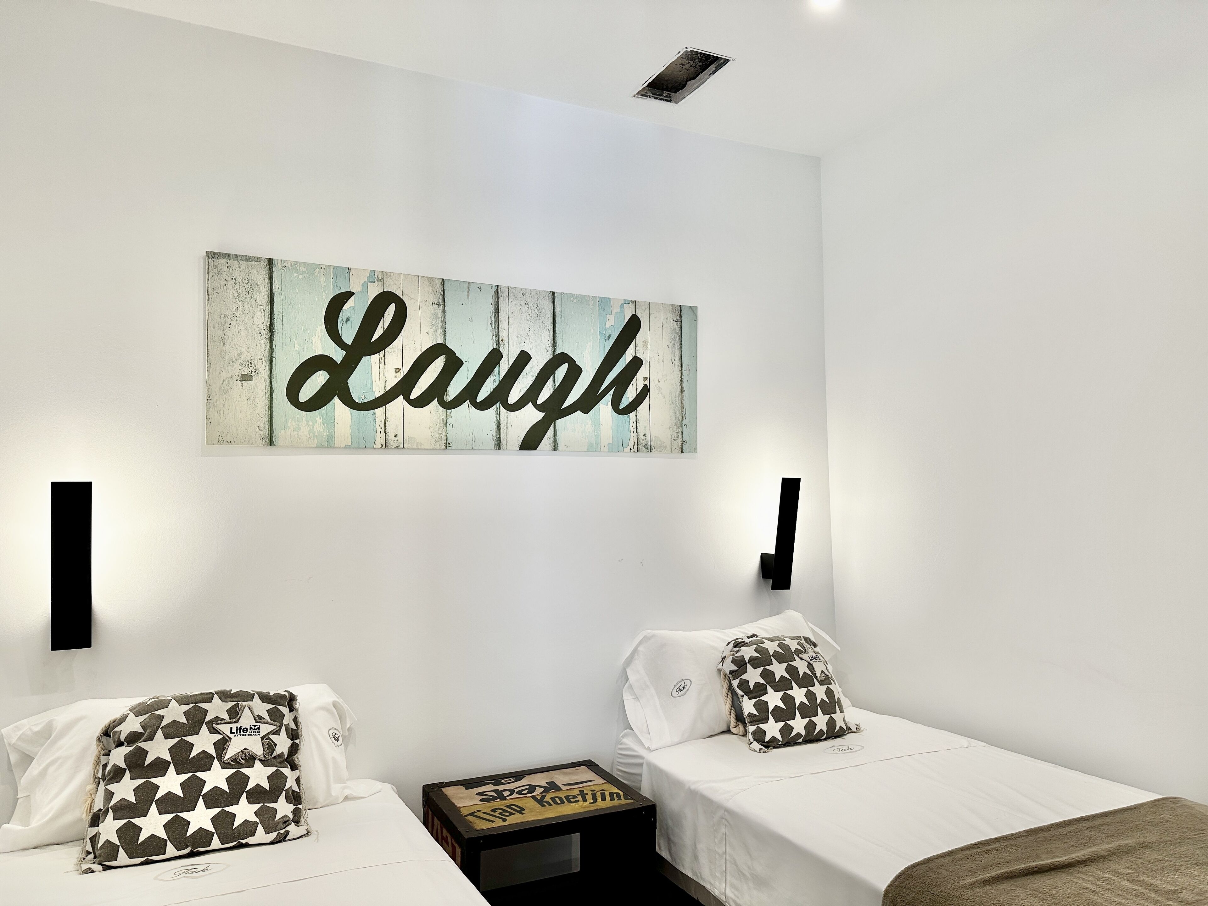 Ein Schlafzimmer mit zwei Betten, jedes mit gemusterten Kissen, einem an der Wand befestigten Schild mit der Aufschrift 'Laugh' und einem kleinen Tisch mit einer Schachtel.