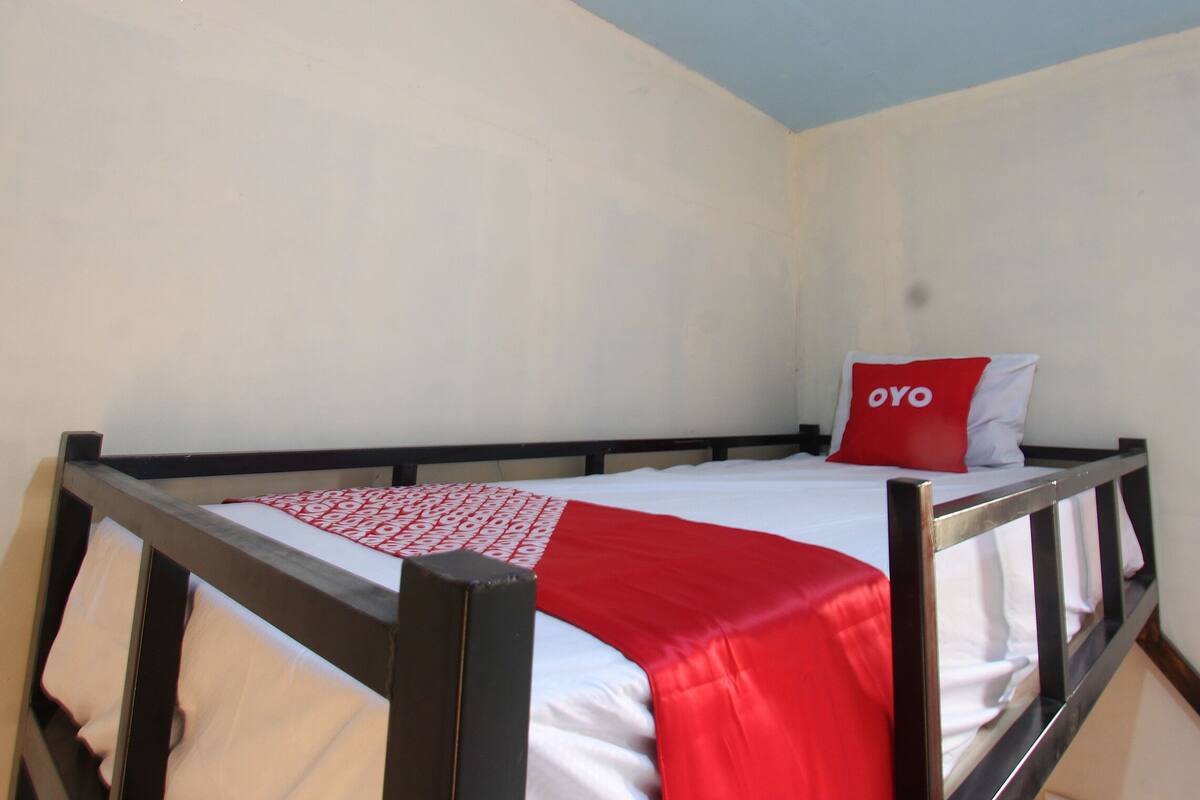 A single bed with a red and white bedspread, a pillow with "OYO" text, and a wooden headboard in a room with plain walls.