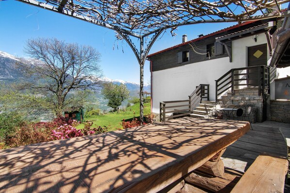 Terraza de madera con vistas a un paisaje montañoso, una escalera de piedra y un edificio con techo rojo.