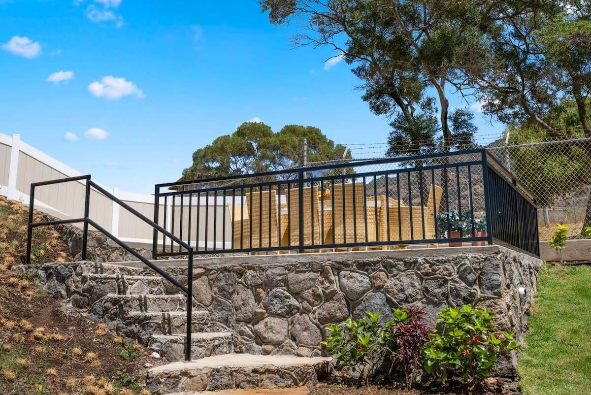 Un escalier en pierre mène à un balcon orné d’une rambarde en métal, le tout étant entouré de verdure et d’arbres.