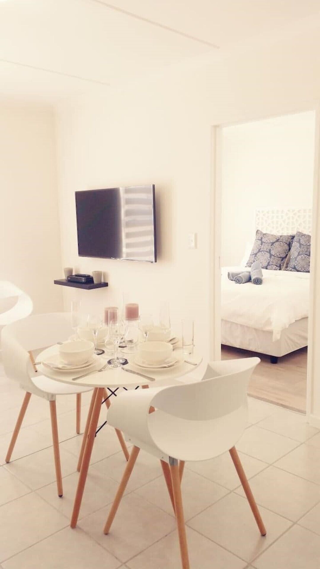 Une chambre d’hôtel moderne dotée d’un coin repas avec une table ronde et des chaises blanches, d  ’un téléviseur à écran plat fixé au mur et d’un lit agrémenté d’oreillers à motifs.