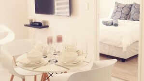 Ett modernt hotellrum med en matplats som har ett runt bord och vita stolar, en platt-TV som är monterad på väggen och en säng med mönstrade kuddar.
