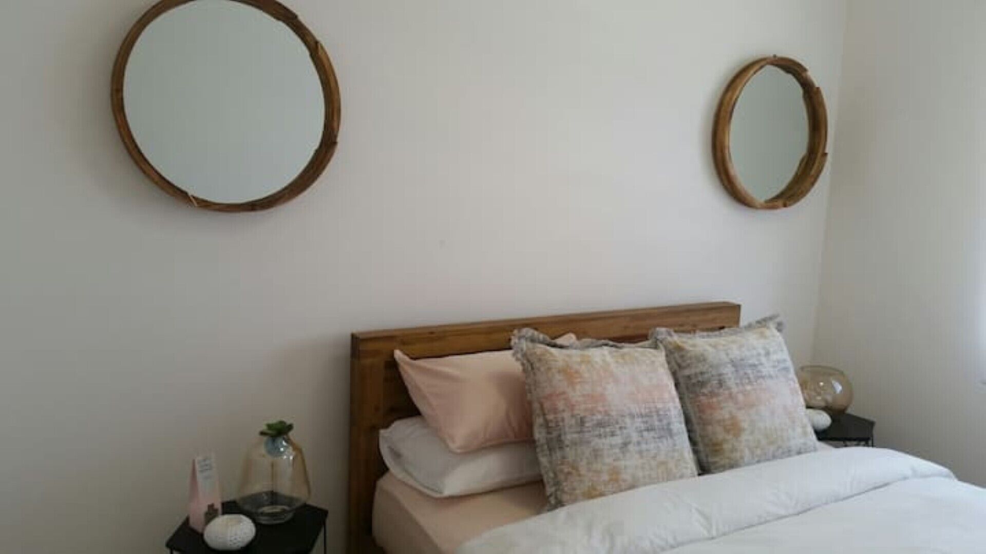 Una camera da letto con una testiera in legno, due comodini, un letto con biancheria da letto bianca e cuscini fantasia, e due specchi fissati alla parete.
