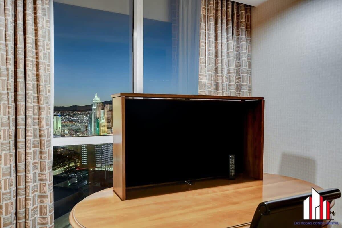 A room with a large window offering a city view, a wooden table, and a remote control on the table.