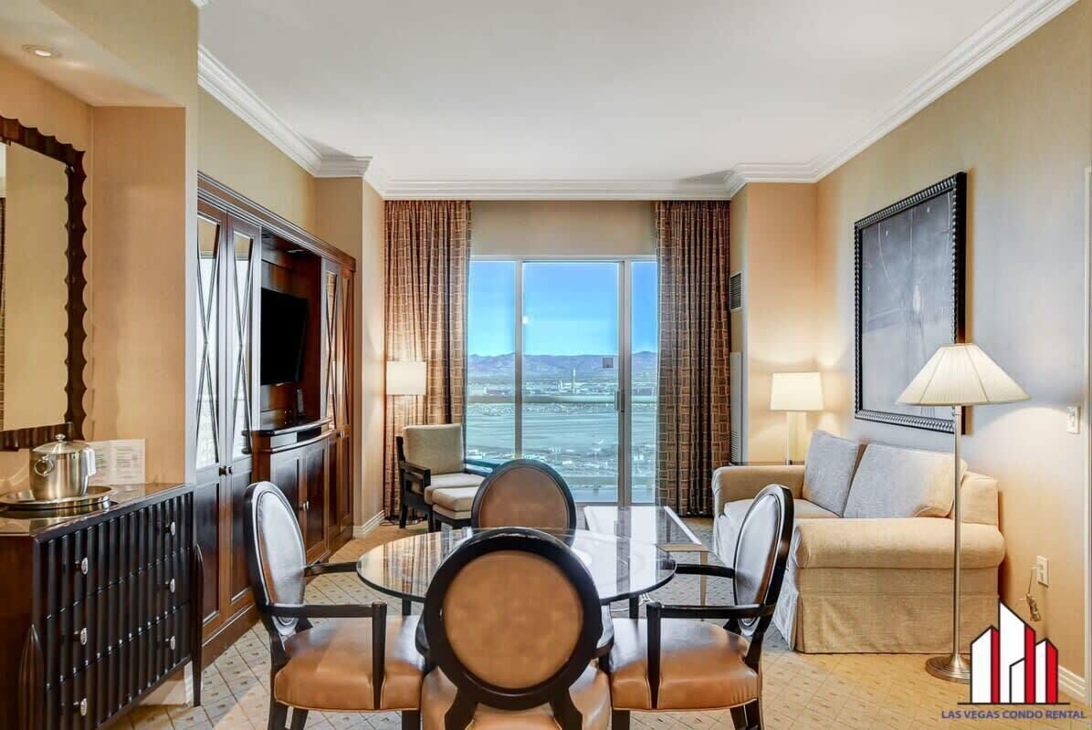A hotel room with a dining area, a sofa, a TV, and a large window with a view.