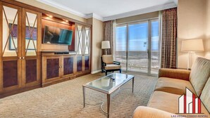 Una sala de estar con un televisor grande, una mesa de centro de vidrio, un sofá y una silla. Hay un ventanal con vistas a la ciudad.