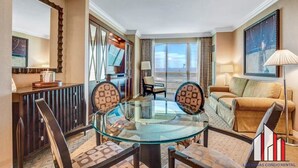 Habitación de hotel con una mesa de vidrio, dos sillas, un sofá, una lámpara y un amplio ventanal con cortinas.