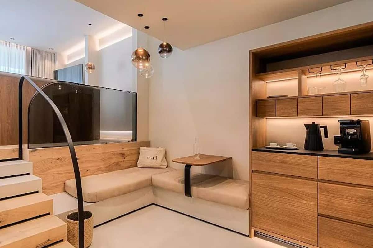 Quarto de hotel moderno com escada de madeira, sofá, mesa pequena e área de cozinha com máquina de café.