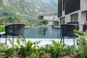 Une piscine avec deux fauteuils en osier au premier plan, un bâtiment moderne doté de balcons en arrière-plan et un paysage montagneux.