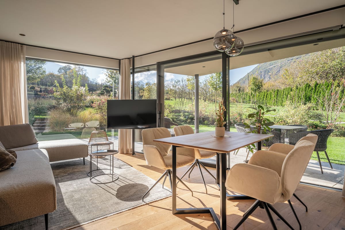 Uma sala de estar moderna com sofá, TV, mesa de jantar com cadeiras e vista para um jardim e montanhas.