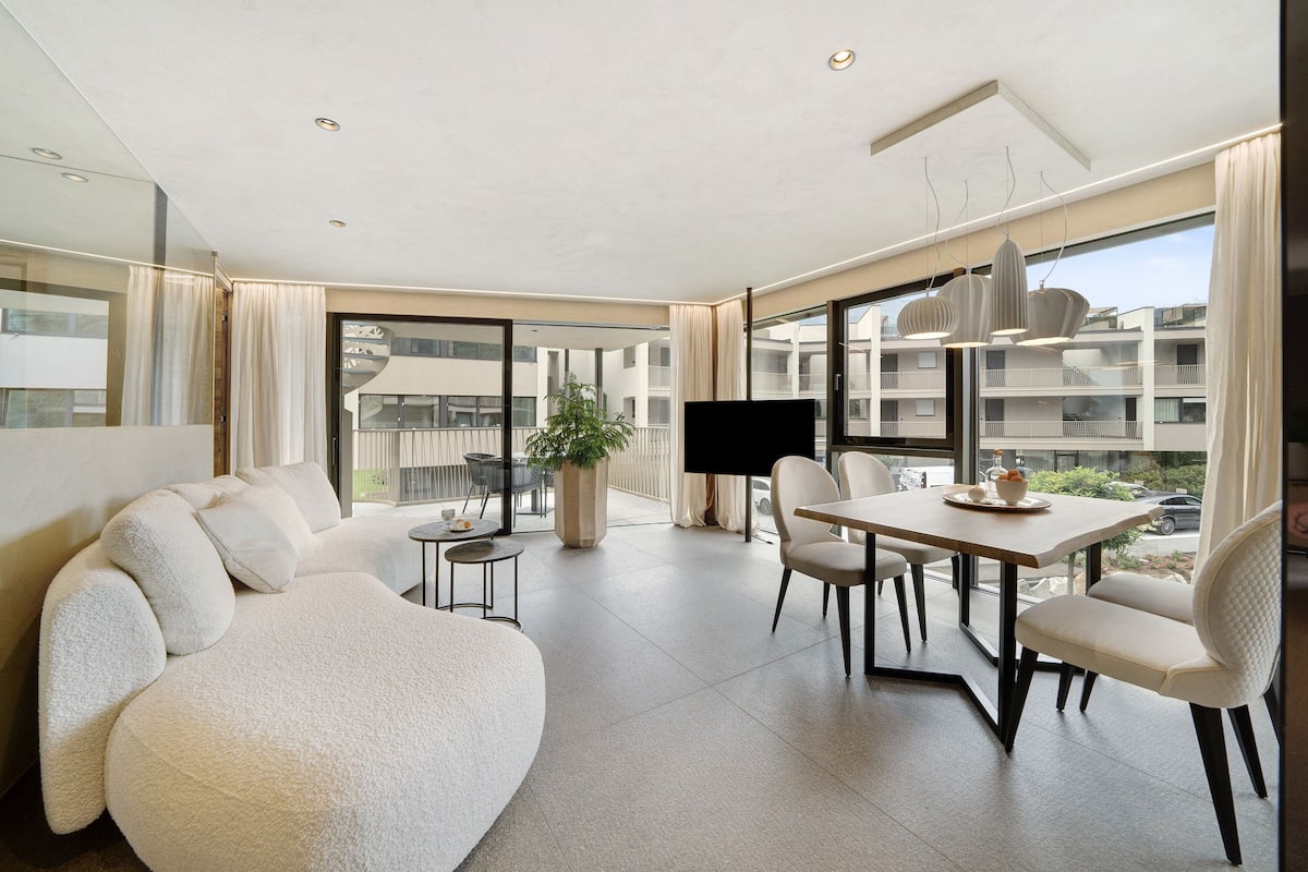 Uma sala de estar moderna com um sofá grande, uma mesa de jantar com cadeiras, uma TV e uma janela ampla com vista para outros edifícios.