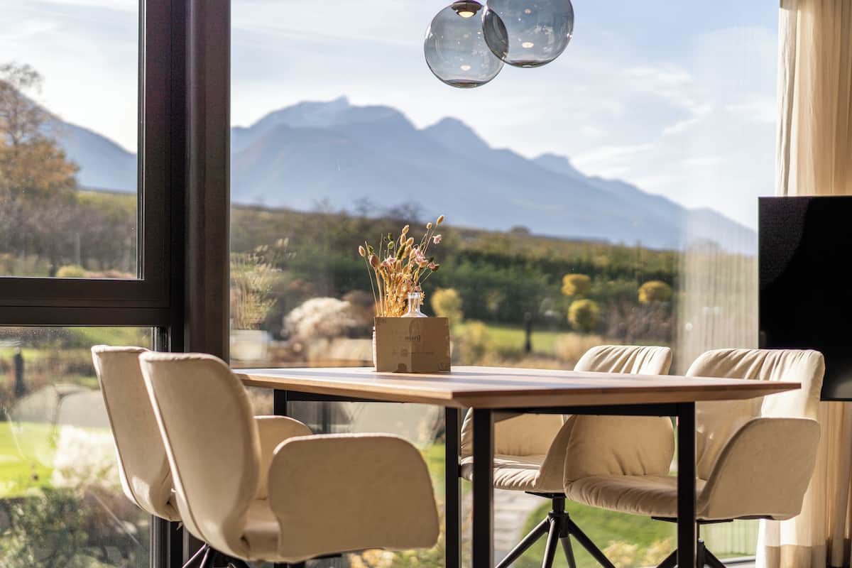 Uma área de refeições com uma mesa e cadeiras, um vaso com flores secas e vista para montanhas através de grandes janelas.