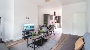 A modern living room with a flat-screen TV, a coffee table, and a sofa. There is a dining area with a table and chairs. The kitchen is visible in the background with cabinets and a countertop. - Spacious 3 Bed Apartment in Waterloo, Liverpool (Liverpool)