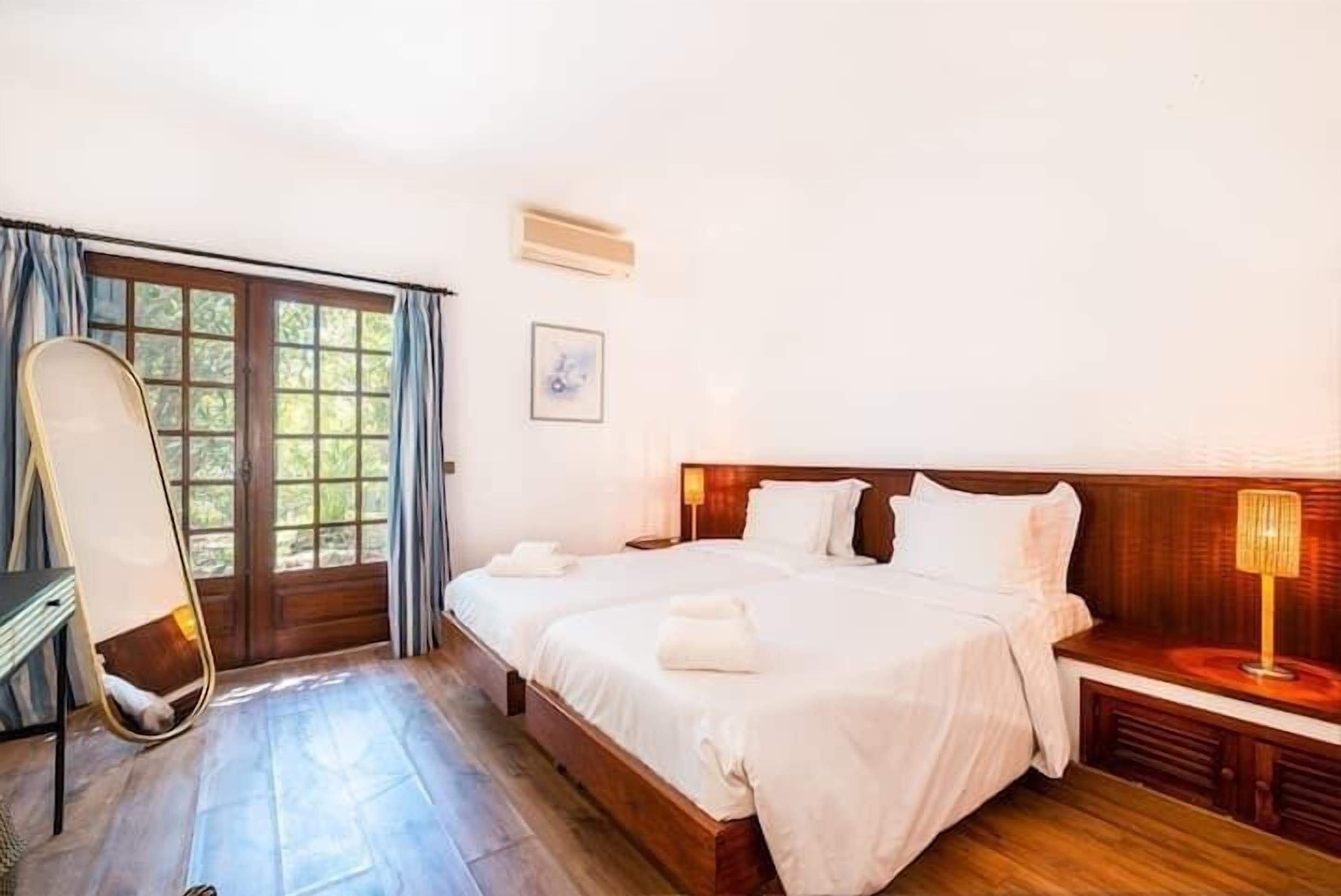 Habitación de hotel con dos camas, cabecero de madera, escritorio con silla, ventana con cortinas y aire acondicionado instalado en la pared.