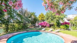 A circular swimming pool with clear water, surrounded by a wooden deck and lush greenery, including pink flowering trees. - Villa Flores - Close to the Amendoeira Golf Resort! (Pêra)