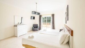 Un dormitorio con cama, cómoda, silla, televisor y ventana con cortinas.