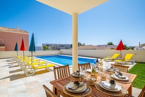 Un área de comedor junto a la piscina con una mesa preparada para cuatro personas, sillones amarillos y sombrillas rojas.
