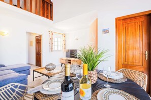 A dining area with a table set for two, featuring a bottle of wine and a glass. There are wicker chairs and a potted plant.