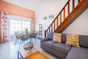 A living room with a grey sofa, a wooden staircase, and a dining area with wicker chairs and a round table.