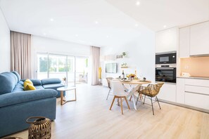 Un salon moderne avec un canapé bleu, une table à manger avec des chaises et un coin cuisine doté de meubles blancs et d’appareils électroménagers.