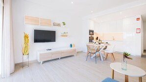 Un salon moderne doté d’un téléviseur à écran plat, d’un coin repas avec une table et des chaises, et d’une cuisine équipée de meubles et d’appareils électroménagers.