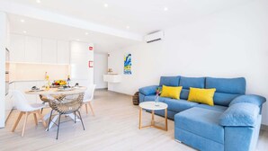 Un salon moderne avec un canapé bleu et des coussins jaunes, un coin repas avec des chaises blanches et une table ronde, et une cuisine avec des meubles blancs.
