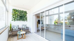 Un salon moderne doté d’une porte coulissante en verre donnant sur un balcon, d’un canapé en osier avec des coussins, d’une petite table sur laquelle est posé un vase, et d’une décoration murale végétale.