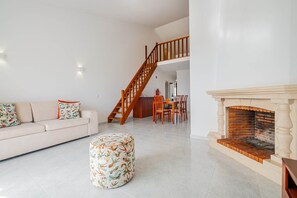 Uma sala de estar com sofá, apoio para os pés, lareira, mesa de jantar, cadeiras e escada.