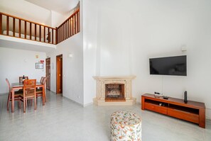 Uma sala de estar com lareira, televisão fixada na parede, mesa de jantar com cadeiras e escada que leva a um andar superior.