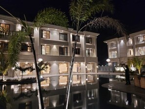 A multi-story hotel with a reflective pool, palm trees, and well-lit balconies. - The Imperial Prime Clarks Inn Suites (Bhilwara)