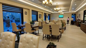 Um restaurante amplo com várias mesas e cadeiras, uma televisão na parede e janelas grandes com vista para o exterior.