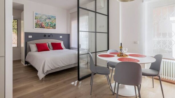 A modern bedroom with a bed, a glass partition, a dining table with chairs, and a pendant light. - Delightful apartment in Ponte Milvio district (Roma)