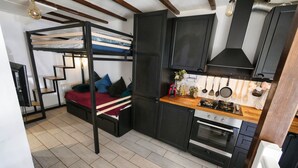 Een compacte keuken met een inbouwfornuis, oven en zwarte kastjes. Een stapelbed met een rode deken staat tegen de muur, en er zijn houten treden die er naartoe leiden.
