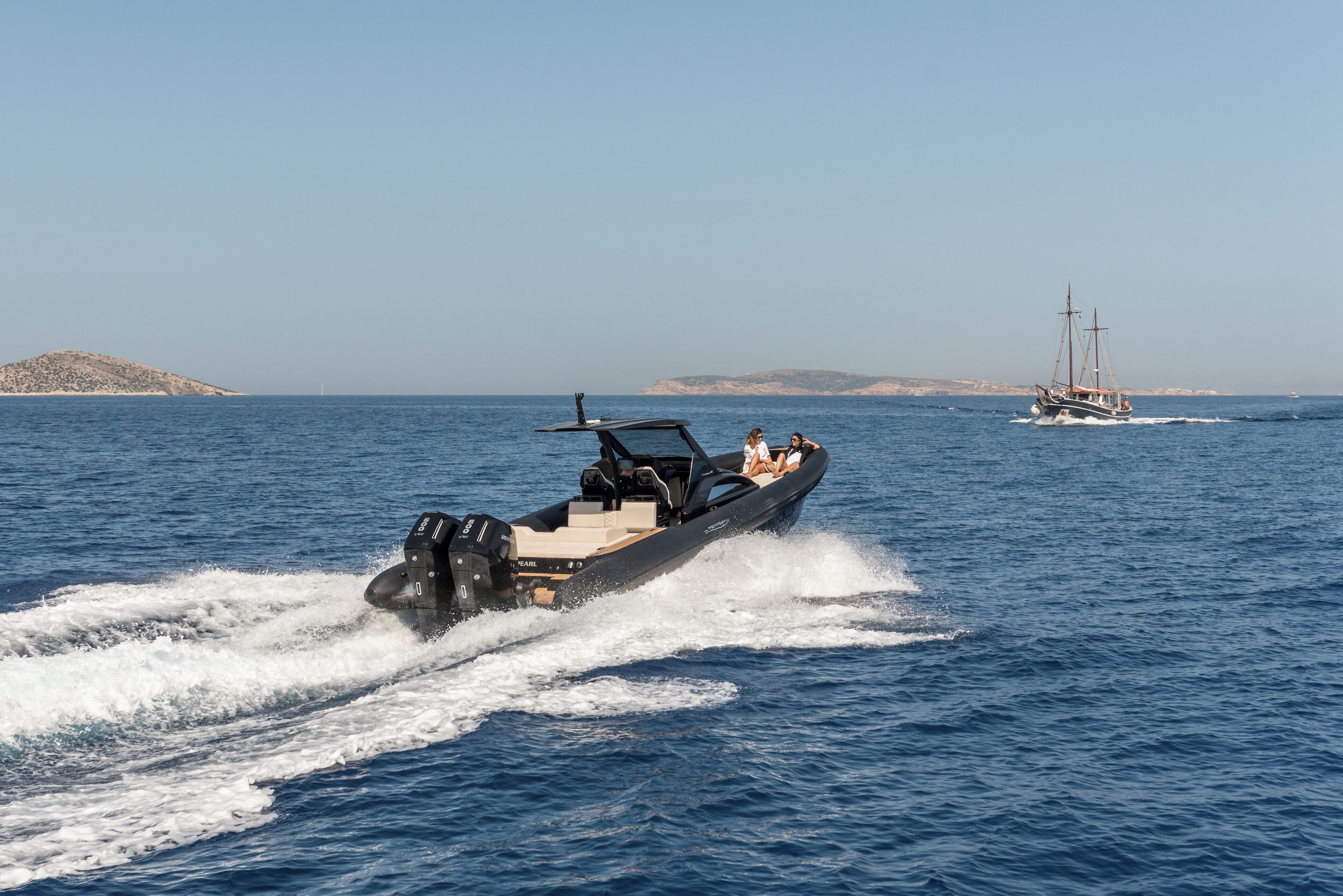 A speedboat with two passengers on a clear day, heading towards a sailboat in the distance.