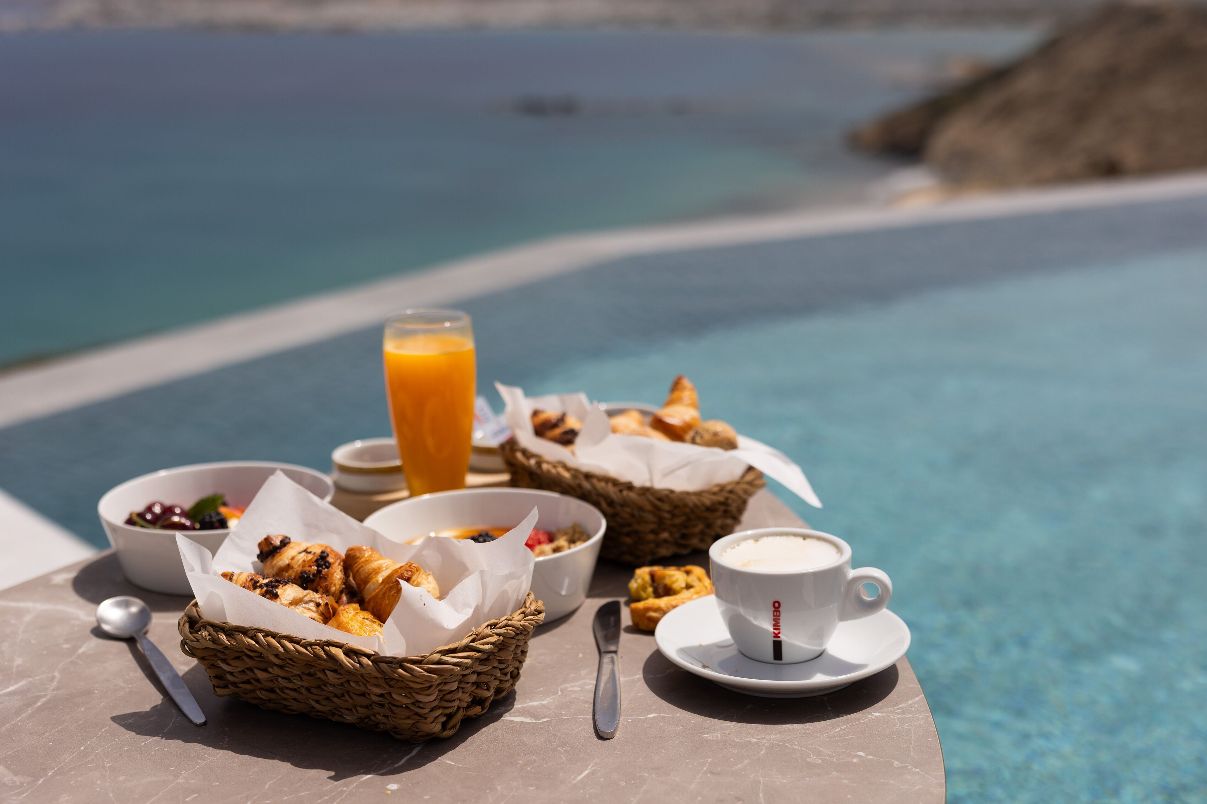 A breakfast setup by a pool with croissants, pastries, a bowl of fruit, a glass of orange juice, and a cup of coffee.