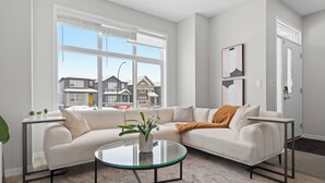 Una sala de estar moderna con un sofá seccional, una mesa de centro de vidrio y vistas a casas a través de amplias ventanas.
