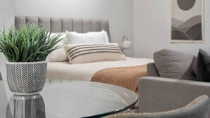 Un dormitorio moderno con una cama, una mesita de noche con planta en maceta, una mesa redonda de cristal y un sofá gris.