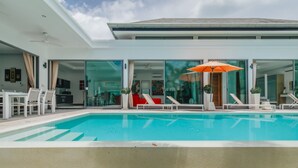 Un área de piscina moderna con agua azul cristalina, sillas reclinables blancas y un paraguas naranja.