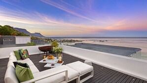 A rooftop terrace with a white sofa, a dining table with fruit and drinks, and a view of the ocean and mountains.