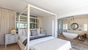 A spacious bedroom with a four-poster bed, a large window with curtains, a bedside table with a lamp, and a view of the bathroom with a bathtub and vanity.
