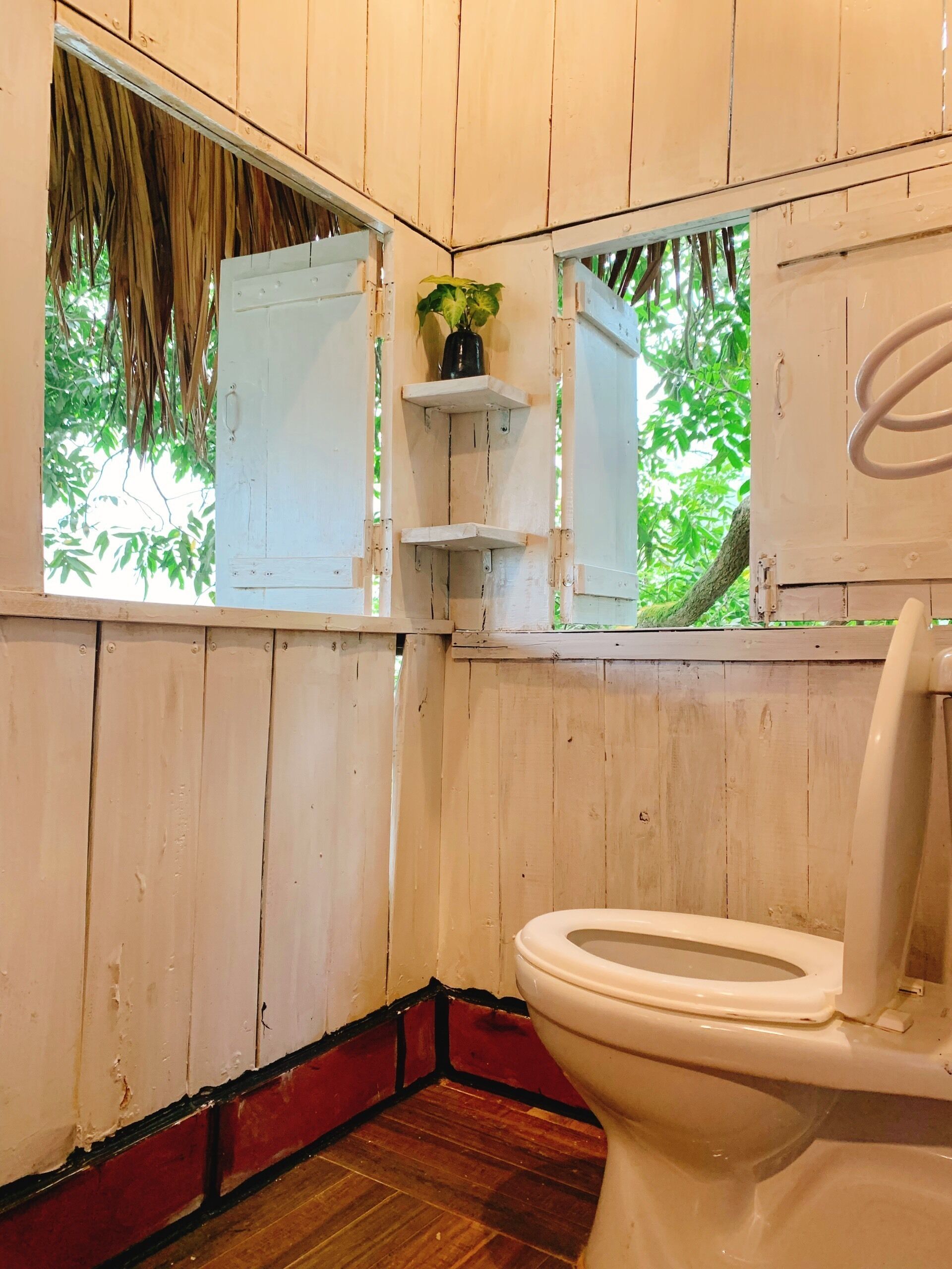 A bathroom with wooden walls, a toilet, a small shelf with a plant, and a window with a view of greenery.