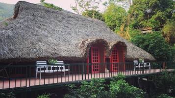 Uma construção com telhado de palha, portas vermelhas, área de estar coberta e guarda-corpo verde.