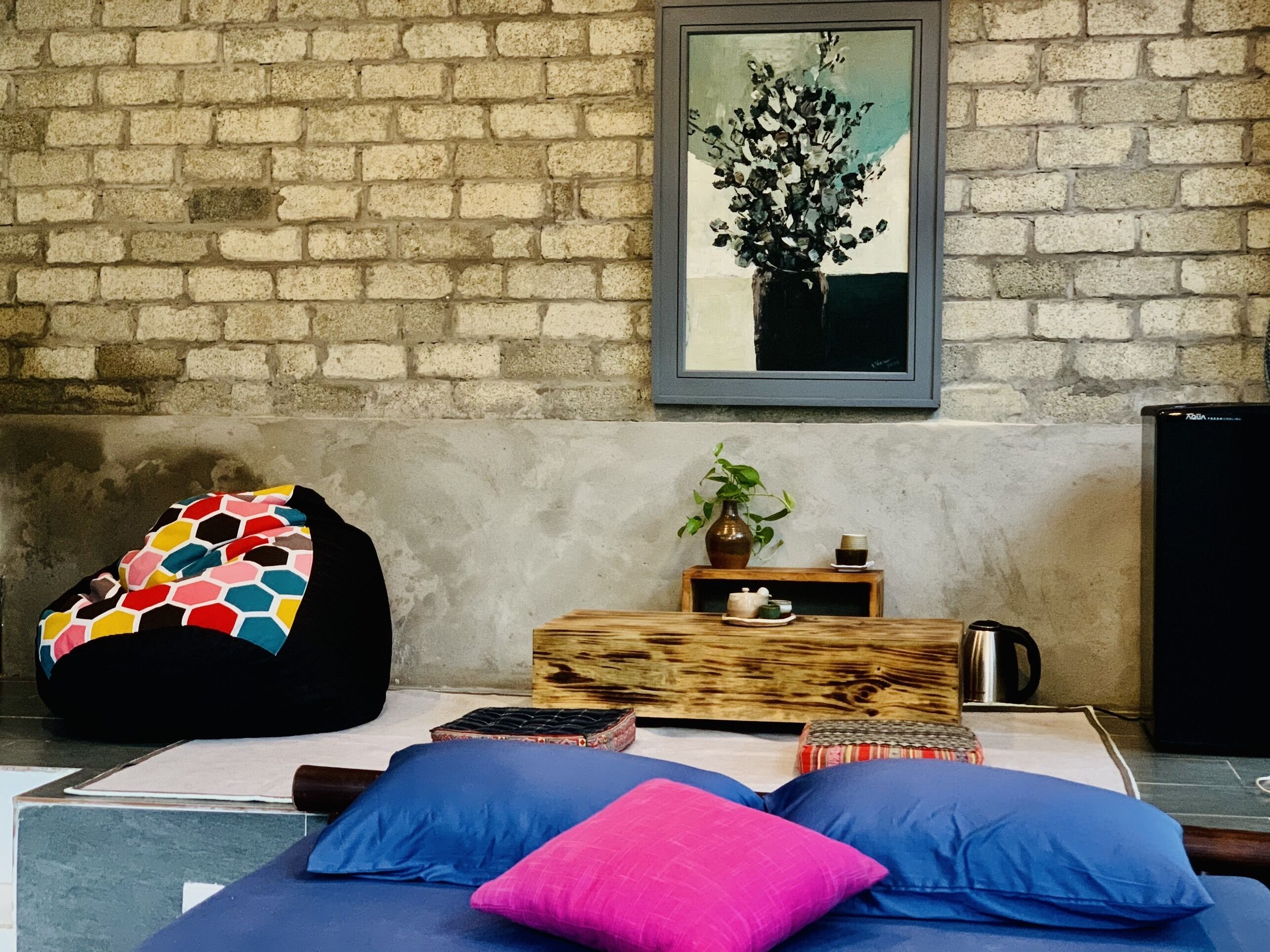 Een kamer met een bakstenen muur, een bed met blauwe en roze kussens, een kleurrijke relaxfauteuil, een klein houten tafeltje met een plant en een kop, en een lijst met een schilderij boven het bed.