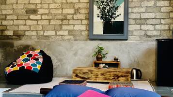 Een kamer met een bakstenen muur, een bed met blauwe en roze kussens, een kleurrijke relaxfauteuil, een klein houten tafeltje met een plant en een kop, en een lijst met een schilderij boven het bed.