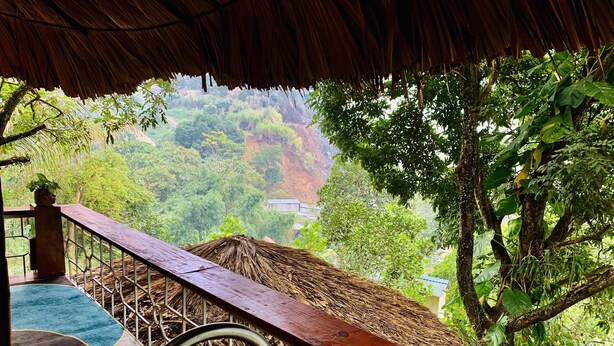 A thatched-roof outdoor area with a wooden bench, a metal railing, and a view of lush greenery and hills in the background. - Muong Retreat (Thong Nhat)