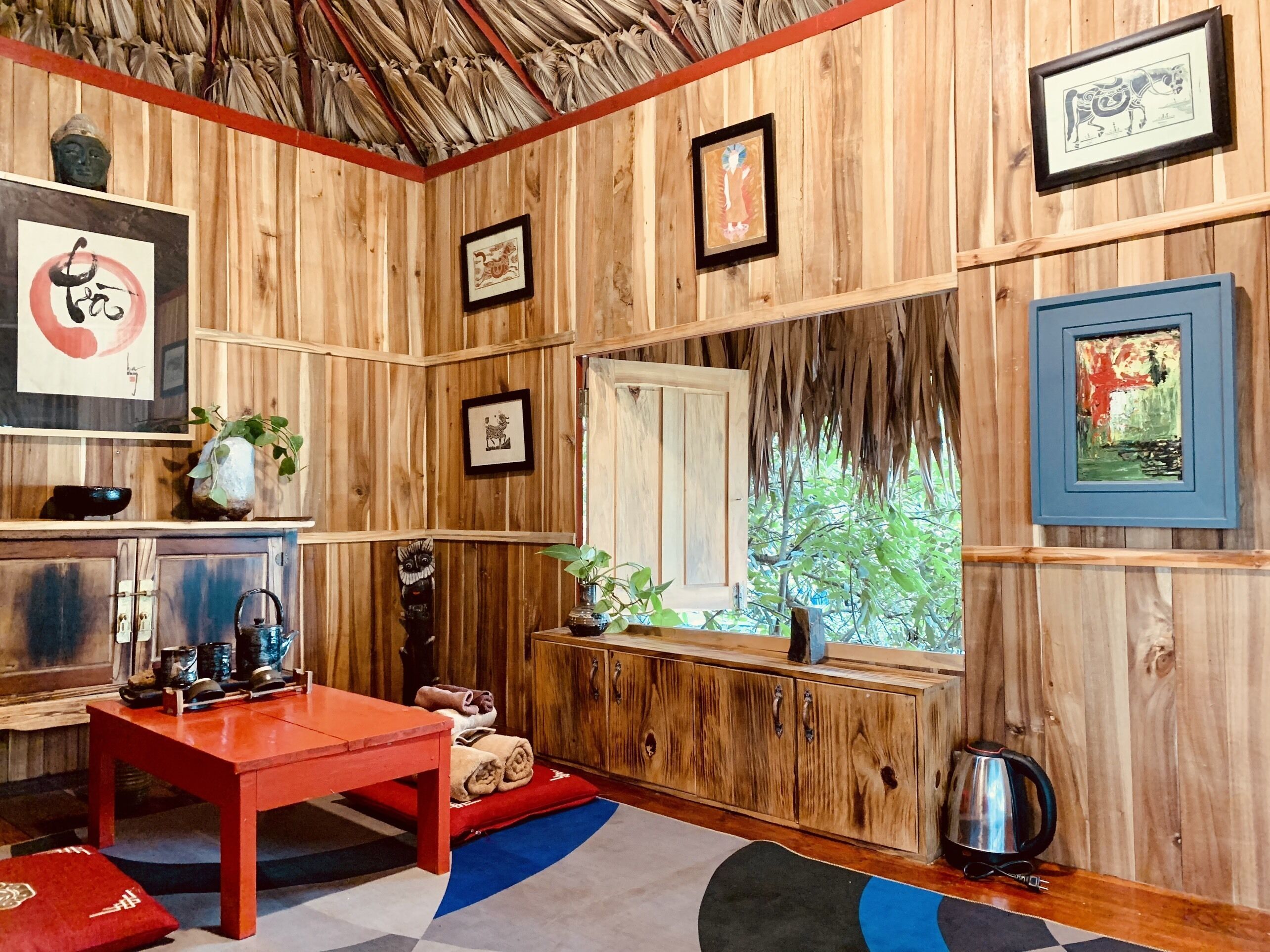 A wooden room with a red table, a window with a view of greenery, and various framed pictures on the walls.