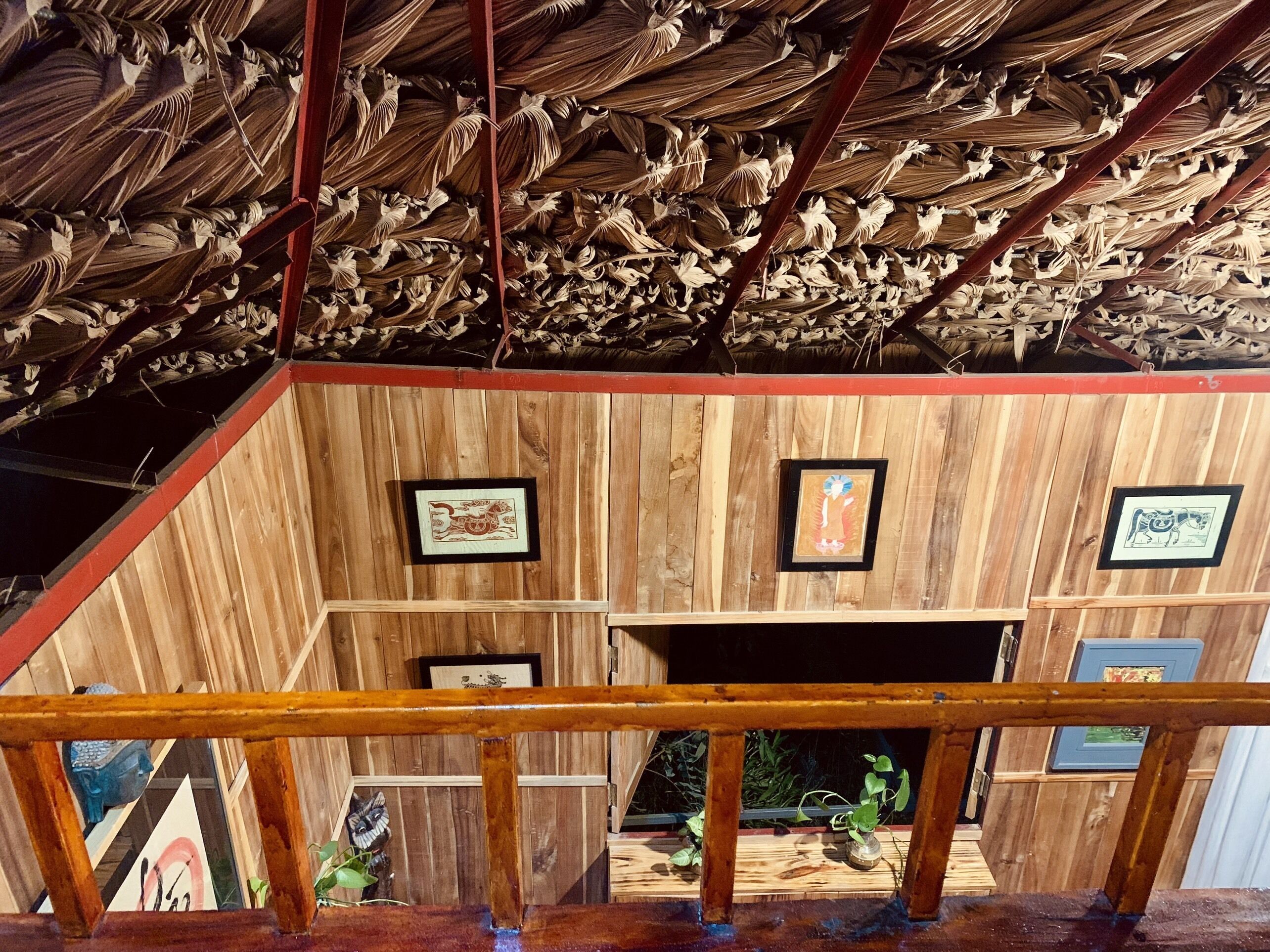 A wooden interior with a sloped ceiling covered in dried leaves, framed artwork on the walls, and a wooden railing in the foreground.