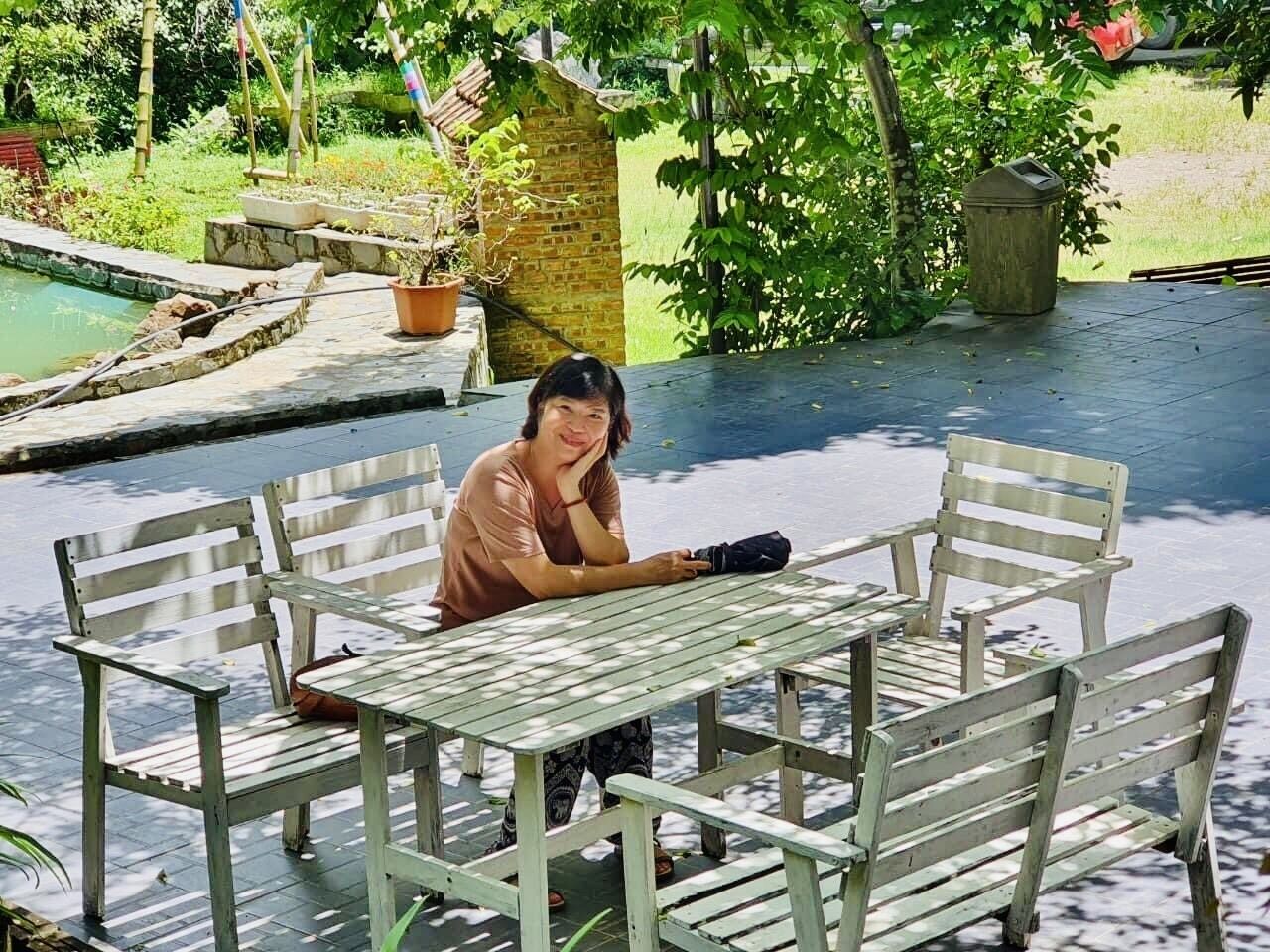En person sitter vid ett träbord med bänkar, utomhus, med en trädgård och en liten pool i bakgrunden.