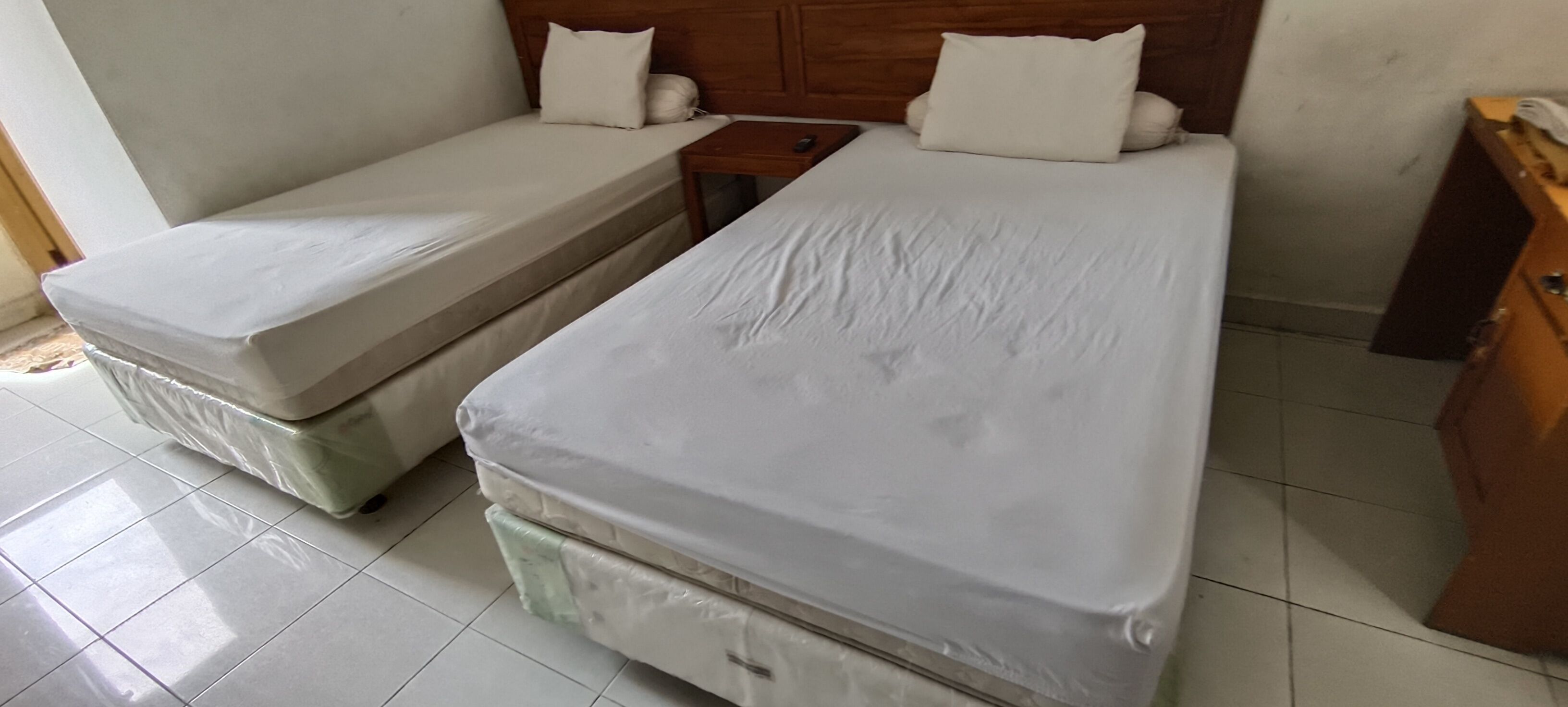 Una habitación con dos camas individuales, cabecero de madera, una mesita de noche pequeña y un armario de madera.